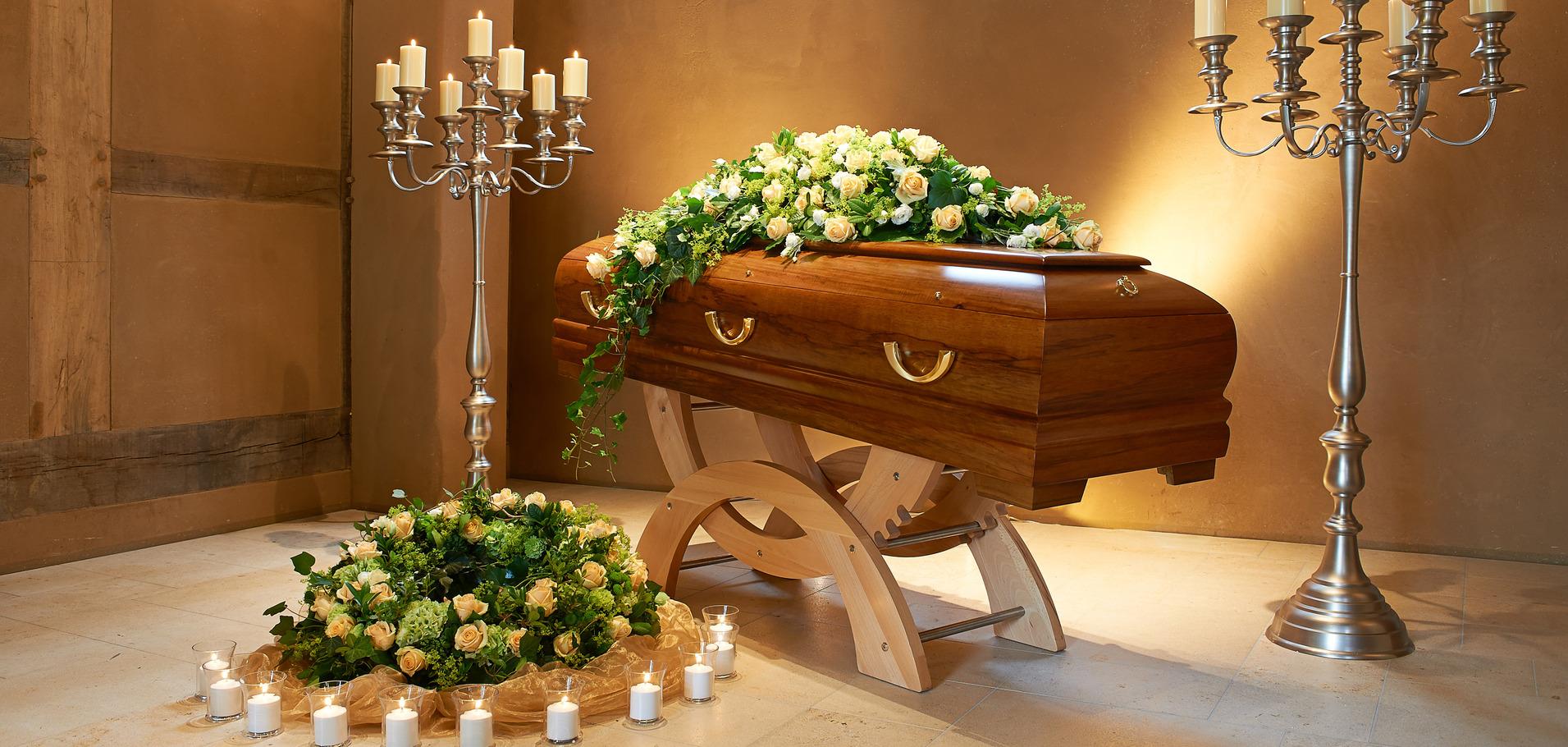 Sarg mit Blumenarrangement in einem Raum, flankiert von Kerzenhaltern und einer Blumendekoration auf dem Boden.