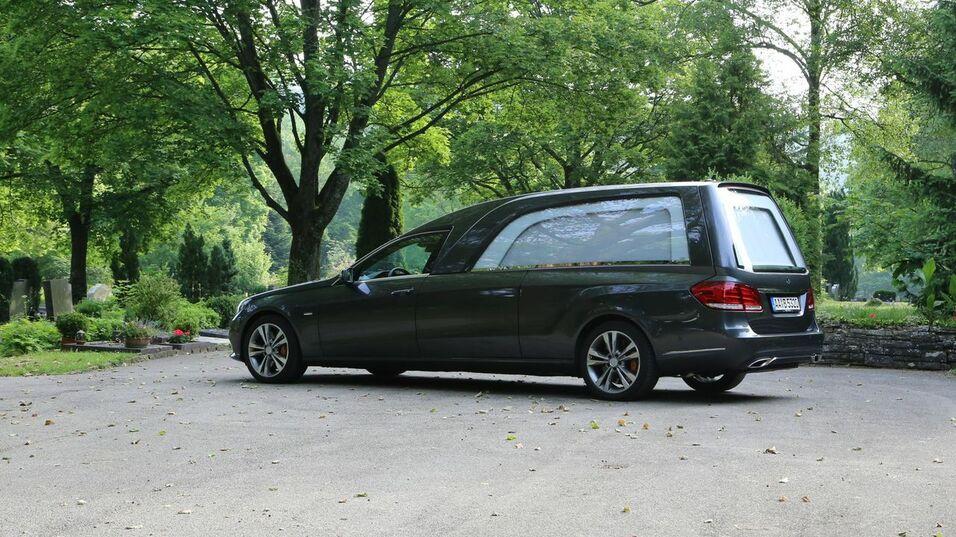 Dunkler Leichenwagen parkt auf einem Weg zwischen Bäumen auf einem Friedhof, neben einer Bank.