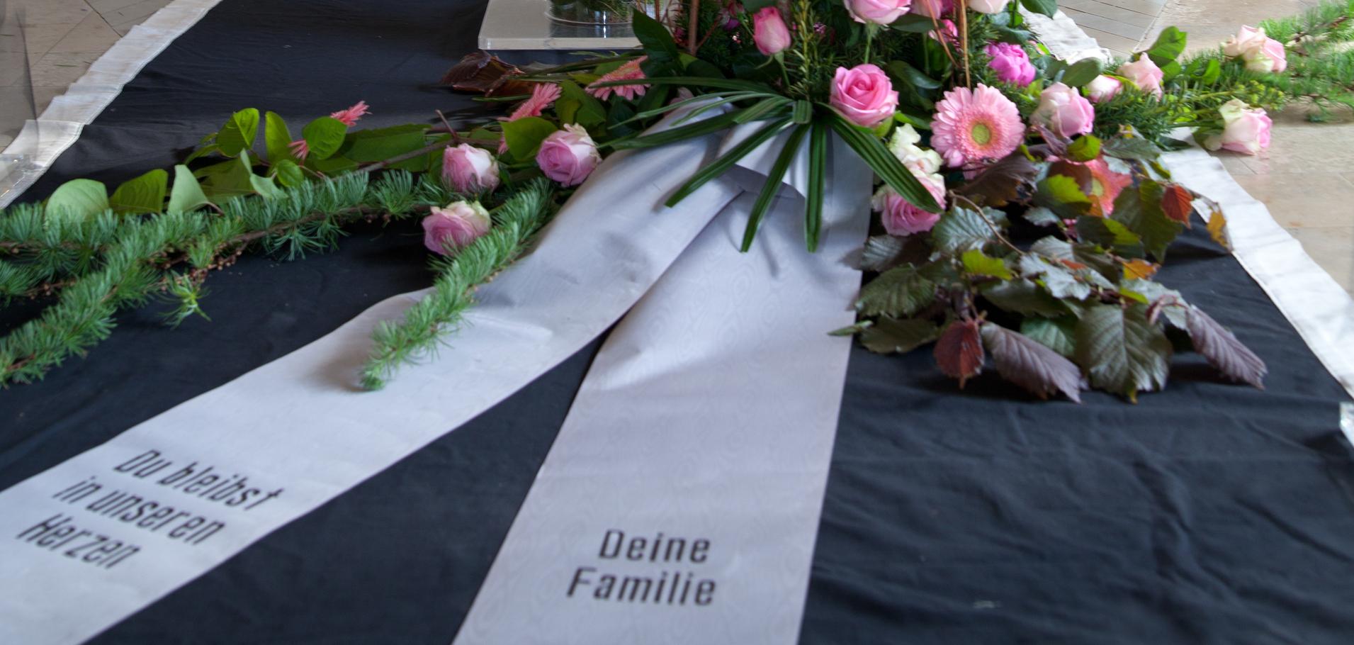 Blumenarrangement mit rosa Rosen und eine Trauerschleife vor einem Kreuz in einer Kapelle.