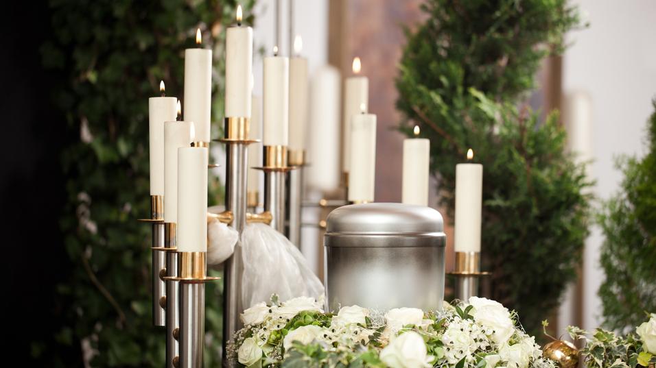 Silberne Urne umgeben von weißen Rosen und Kerzen auf einem Tisch, im Hintergrund grüne Pflanzen.