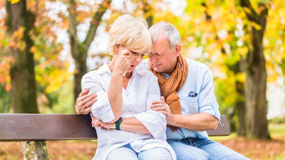 Ein älteres Paar sitzt auf einer Bank im Herbst, mit tröstender Körpersprache und ernsten Gesichtsausdrücken.