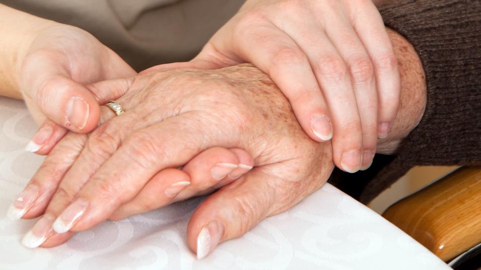 Zwei Hände, eine ältere und eine jüngere, liegen auf einem Tisch, wobei die jüngere Hand die ältere sanft umschließt.