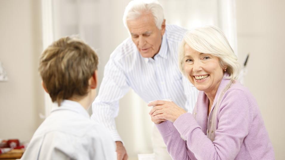 Lächelnde ältere Frau sitzt am Tisch, während ein Mann im Hintergrund Getränke serviert, ein Junge schaut sie an.