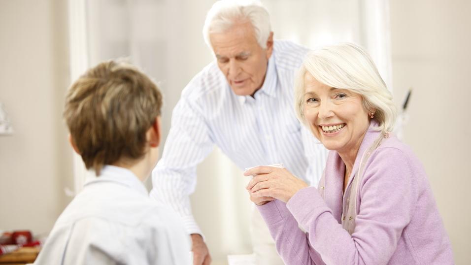 Lächelnde ältere Frau sitzt am Tisch, während ein Mann im Hintergrund Getränke serviert, ein Junge schaut sie an.